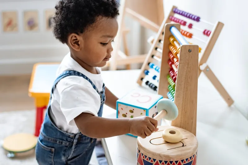 A focused young boy in denim overalls playing with a rainbow wooden abacus and drum