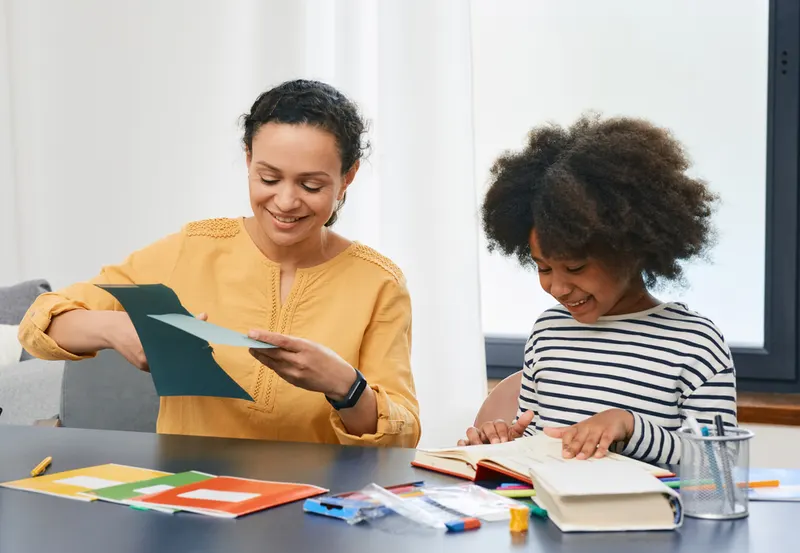 A smiling mother and her young daughter sitting at a table working with books and colorful paper